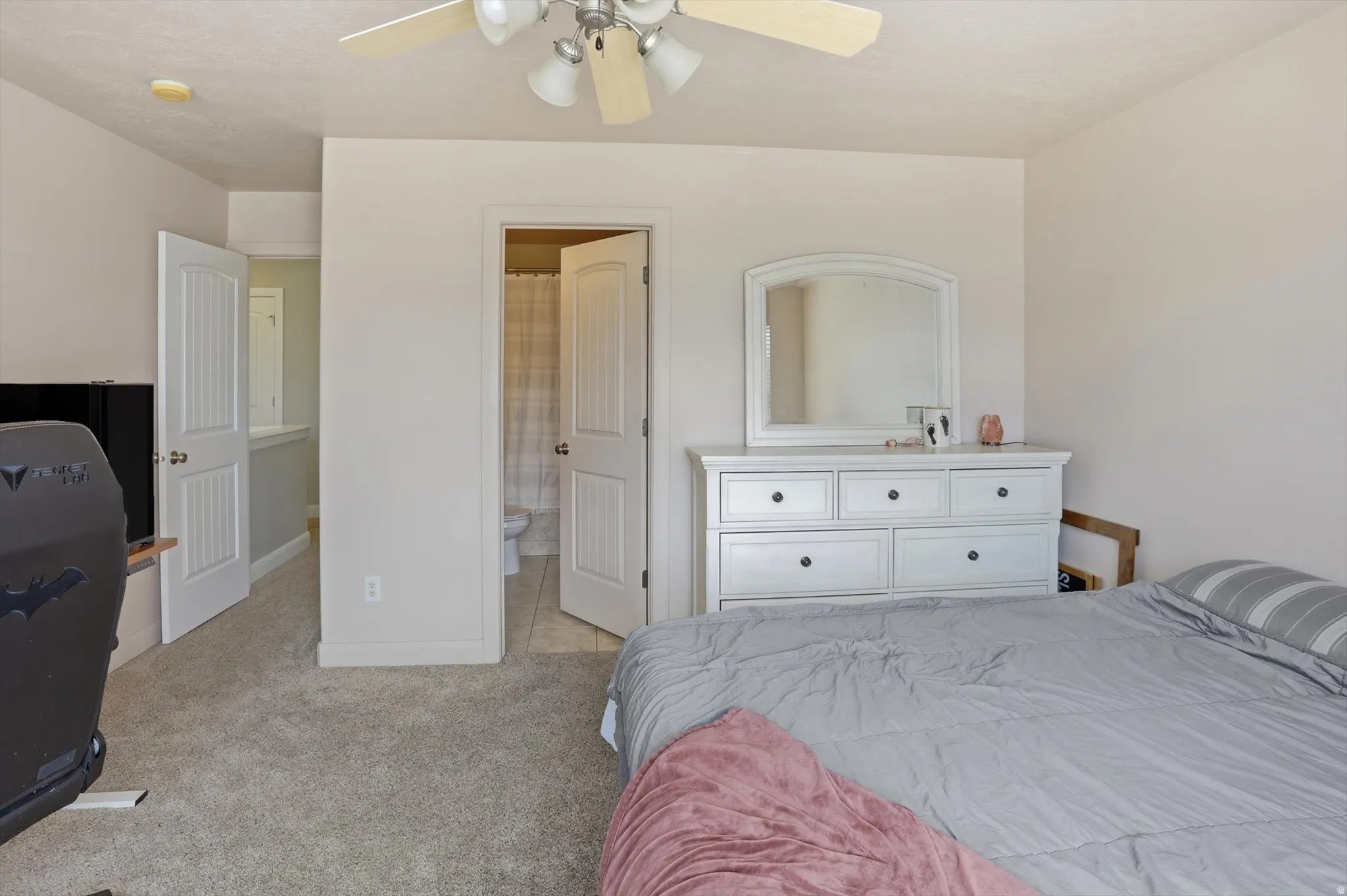 Carpeted bedroom featuring a ceiling fan with light fixture, neutral wall color, and a door opening to a bathroom with tile flooring and a shower curtain
