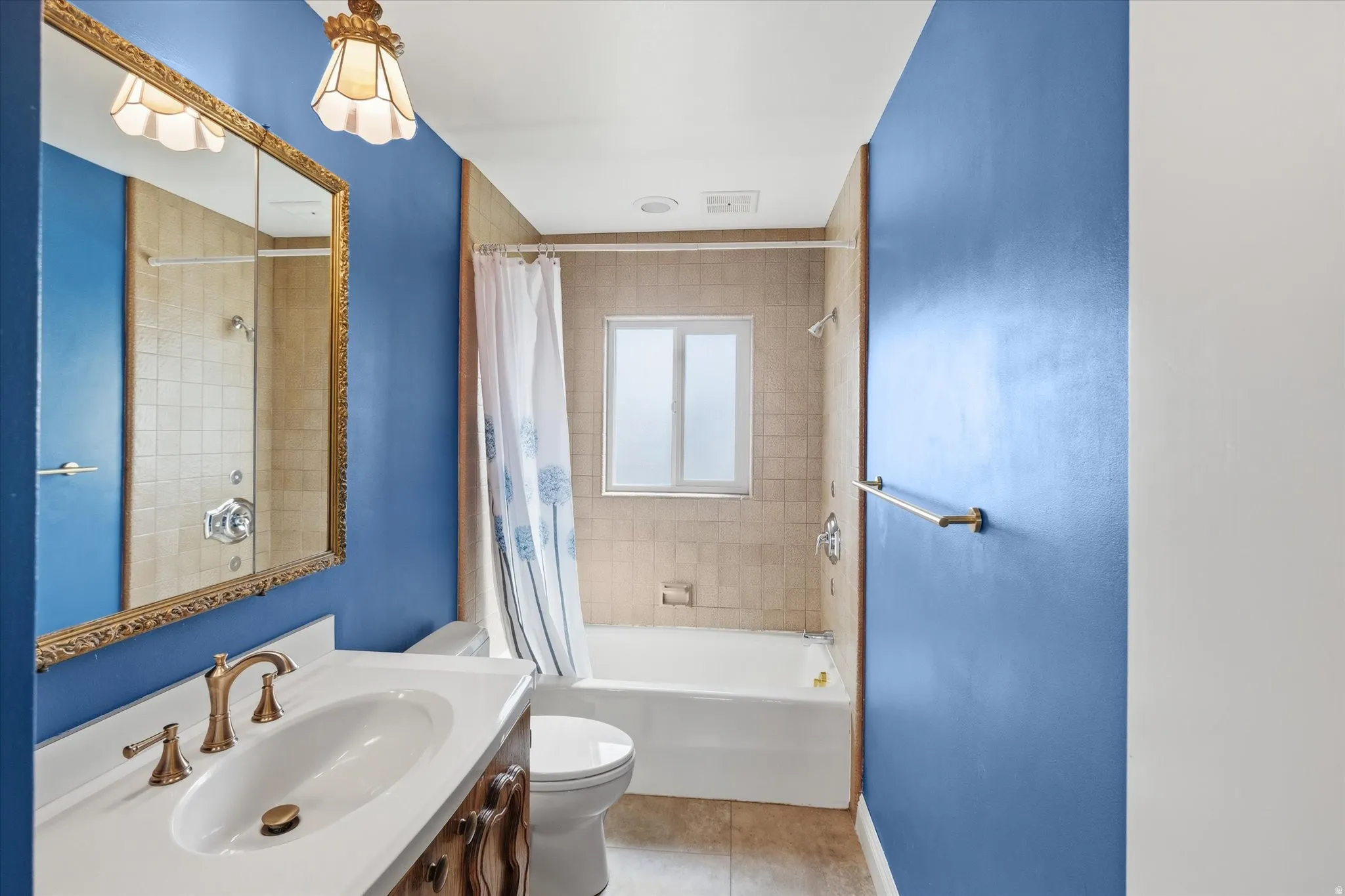 Full bathroom featuring a single vanity with a white countertop and brass-finish faucet, a framed mirror, a tub/shower combination with tan square tiling, and a window