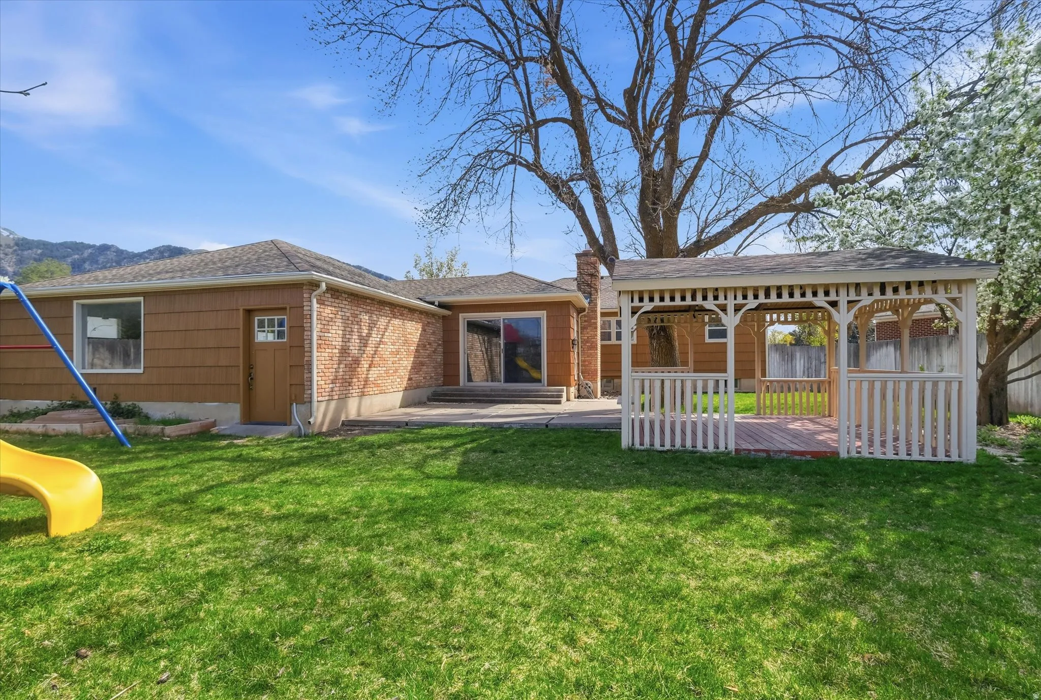 Expansive backyard featuring a white wooden gazebo with a red brick floor, a concrete patio, and a brown siding exterior with brick accents