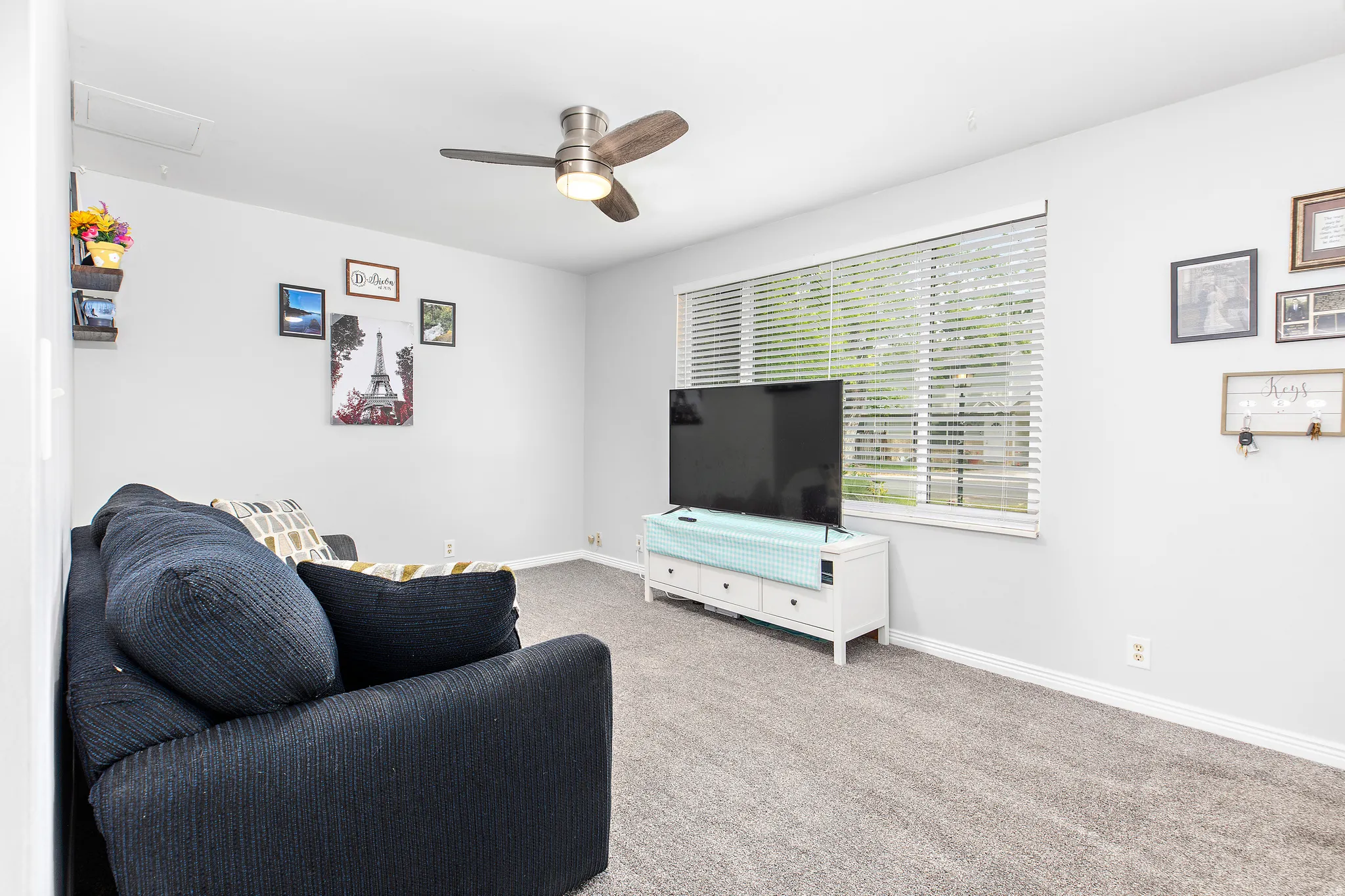 Carpeted living area featuring a ceiling fan with integrated lighting, a large window with horizontal blinds, and neutral wall paint
