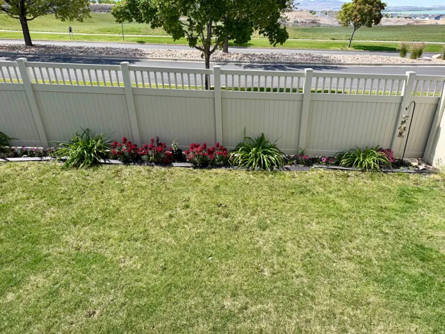 Expansive lawn with a perimeter privacy fence, featuring a landscaped planting bed with mature shrubs and blooming red and pink flowers