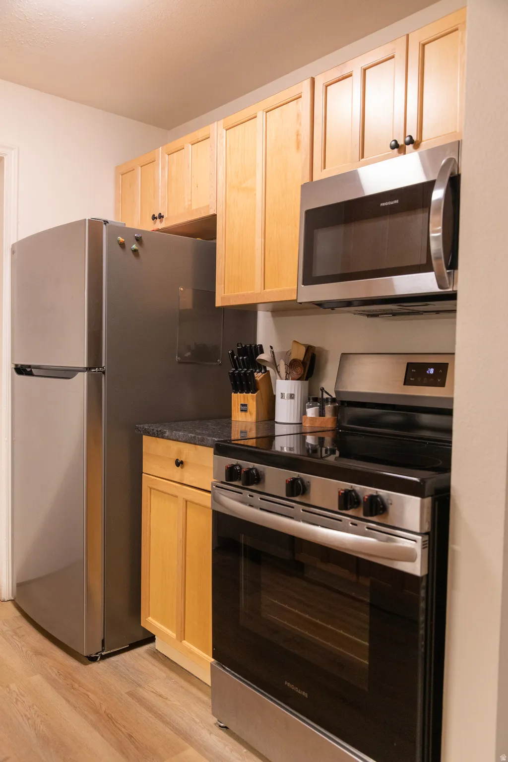 Kitchen featuring light wood cabinetry, stainless steel appliances, dark countertops, and wood-finish flooring