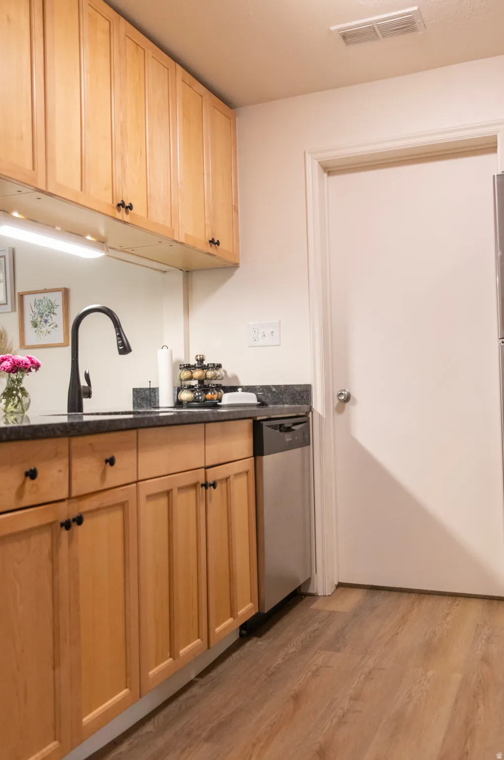 Kitchen featuring light wood cabinetry with black hardware, dark stone countertops, a stainless steel dishwasher, and wood-finish flooring