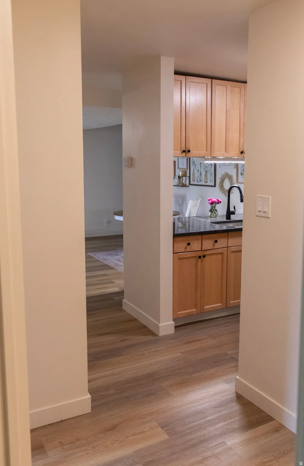 Hallway with wood-finish flooring leading to a kitchen featuring light wood cabinetry, dark countertops, and a black gooseneck faucet