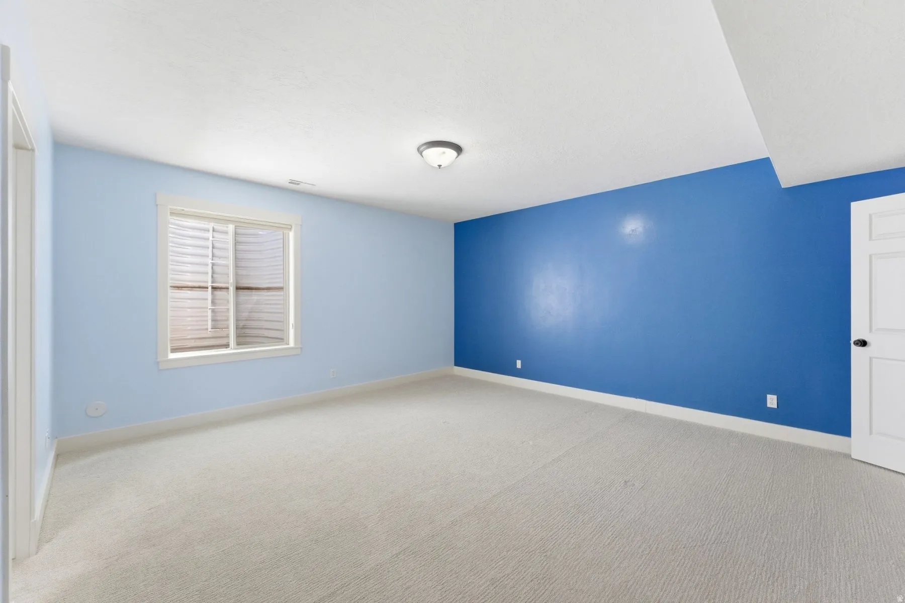 Spacious room featuring light-colored carpeting, a bright window with blinds, and contrasting blue walls