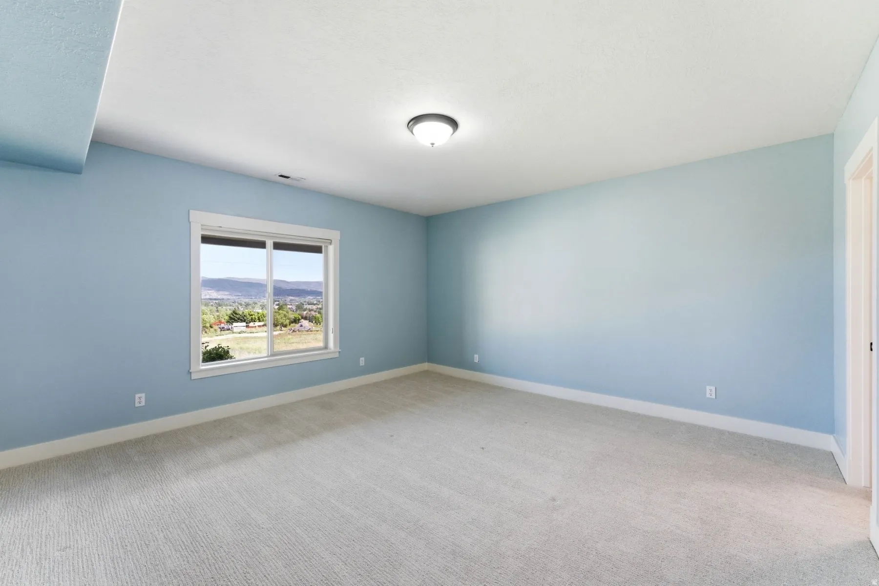 Spacious room with light blue walls, soft neutral carpeting, and a large window offering expansive distant scenery