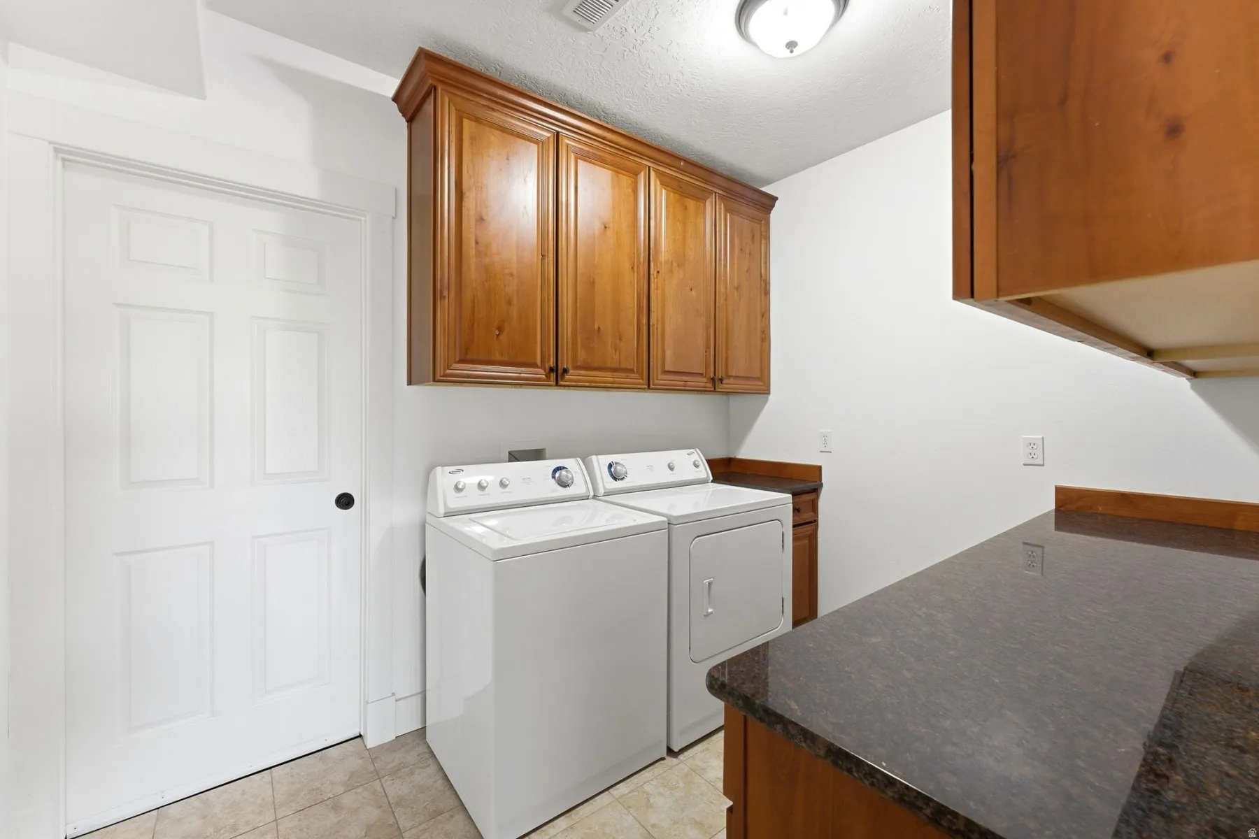 Dedicated laundry space featuring rich wood cabinetry, a dark countertop, overhead lighting, and durable tile flooring