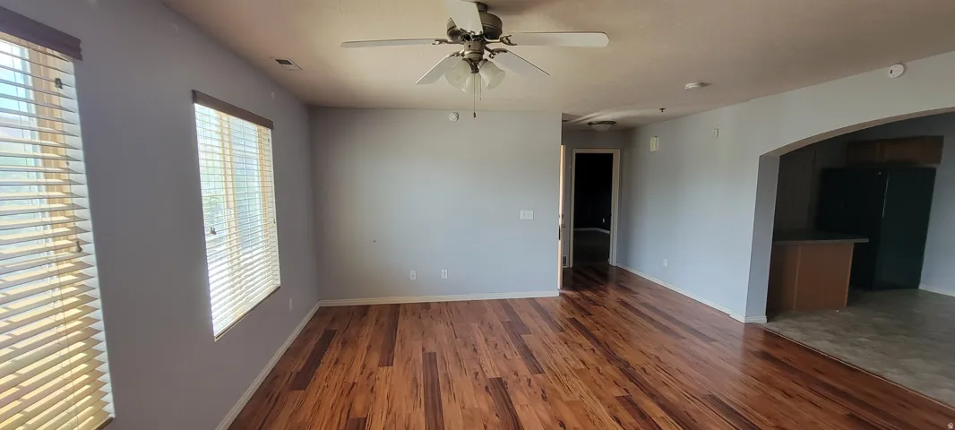 Spacious room featuring wood-finish flooring, two windows with blinds, a ceiling fan with integrated lighting, and an arched entryway