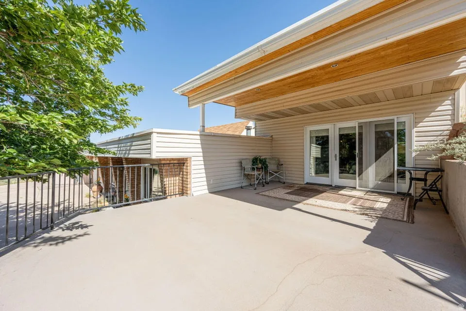 Spacious outdoor concrete patio featuring a partial overhead covering with wood-finish accents, a metal railing, and multiple glass door entries