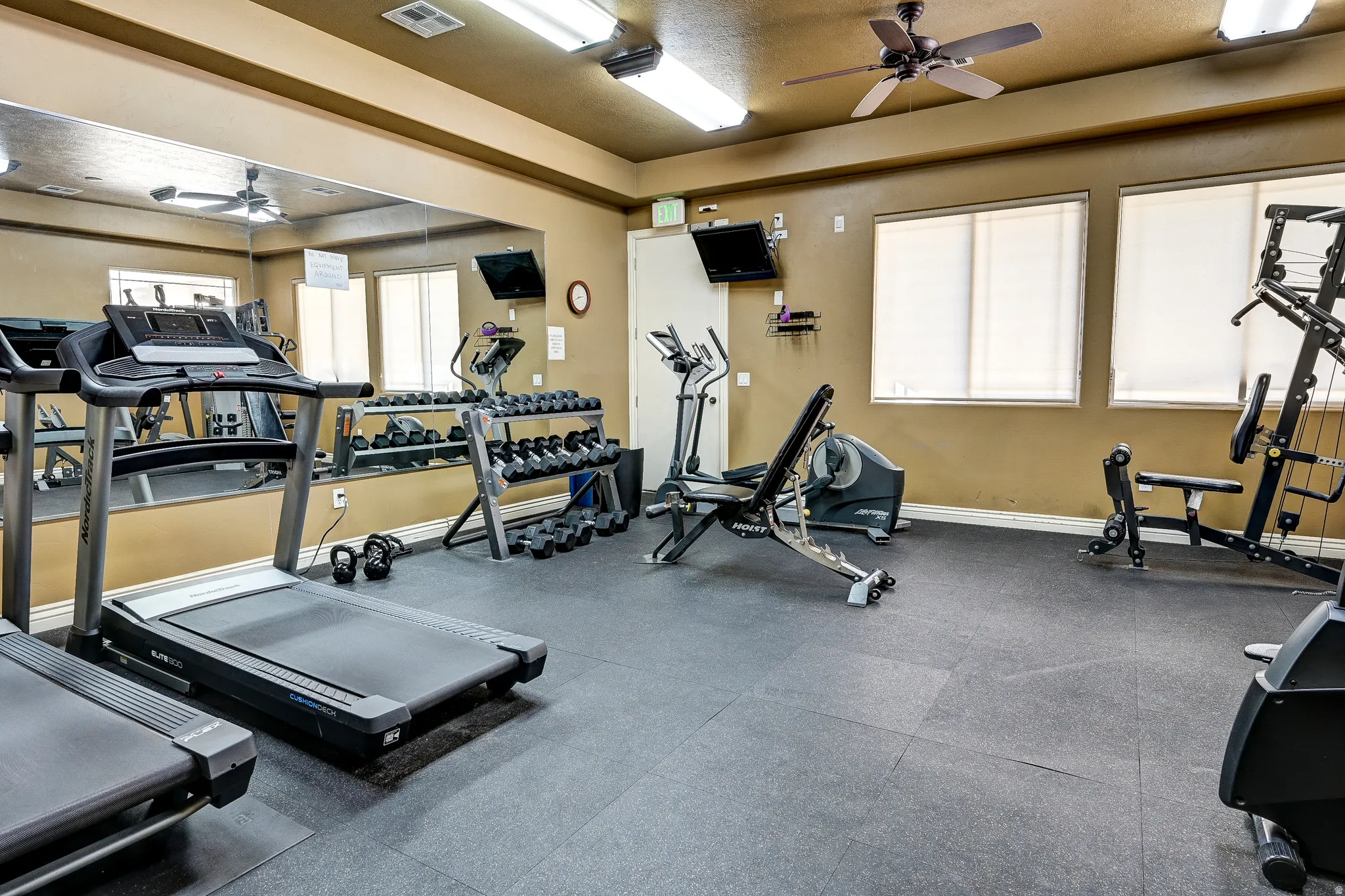 Dedicated fitness room featuring durable rubber flooring, mirrored wall, multiple windows with blinds, ceiling-mounted light fixtures, and a ceiling fan