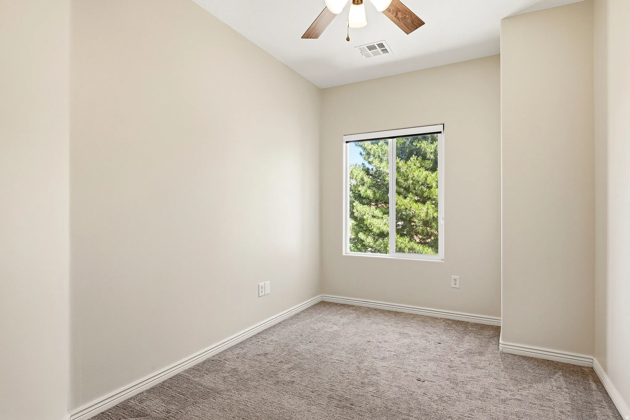 Neutral-toned interior room featuring a window with green tree scenery, a ceiling fan with integrated lighting, carpeted flooring, and white baseboards
