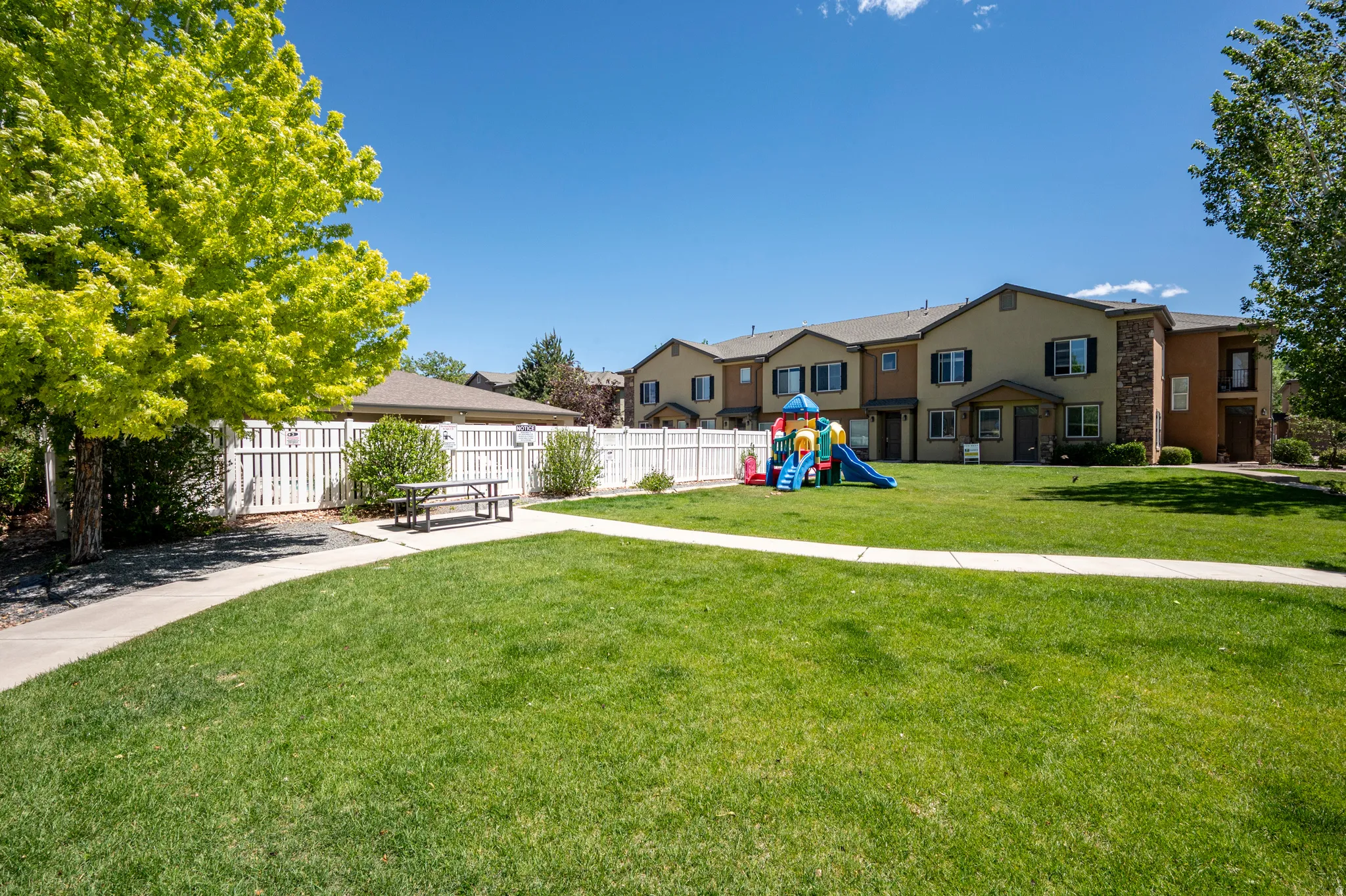 Expansive green space featuring a concrete pathway, a white picket fence, and mature trees