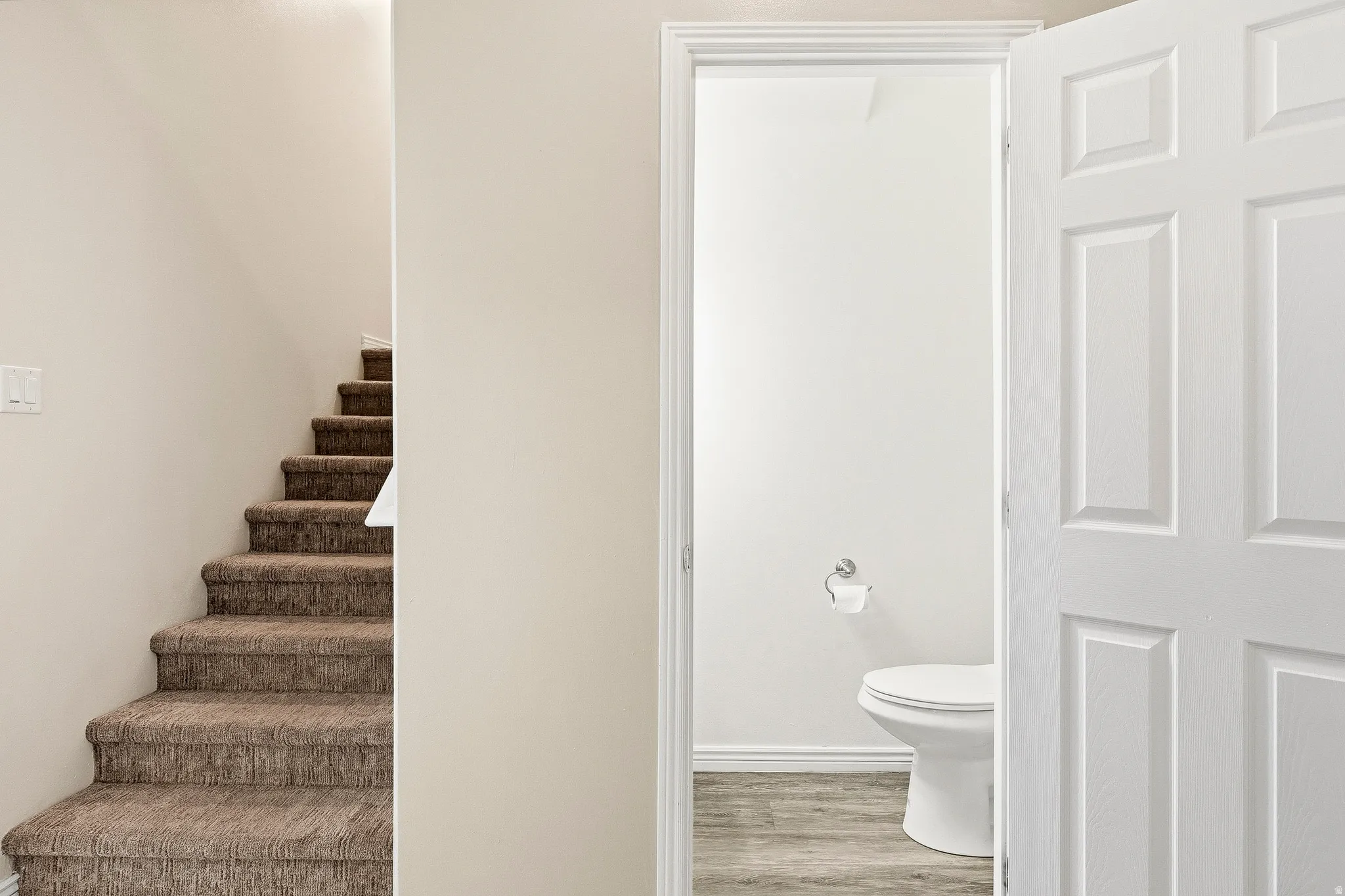 Carpeted stairway with a white light switch and a partial view of a bathroom featuring wood-finish flooring, white baseboards, and a white toilet