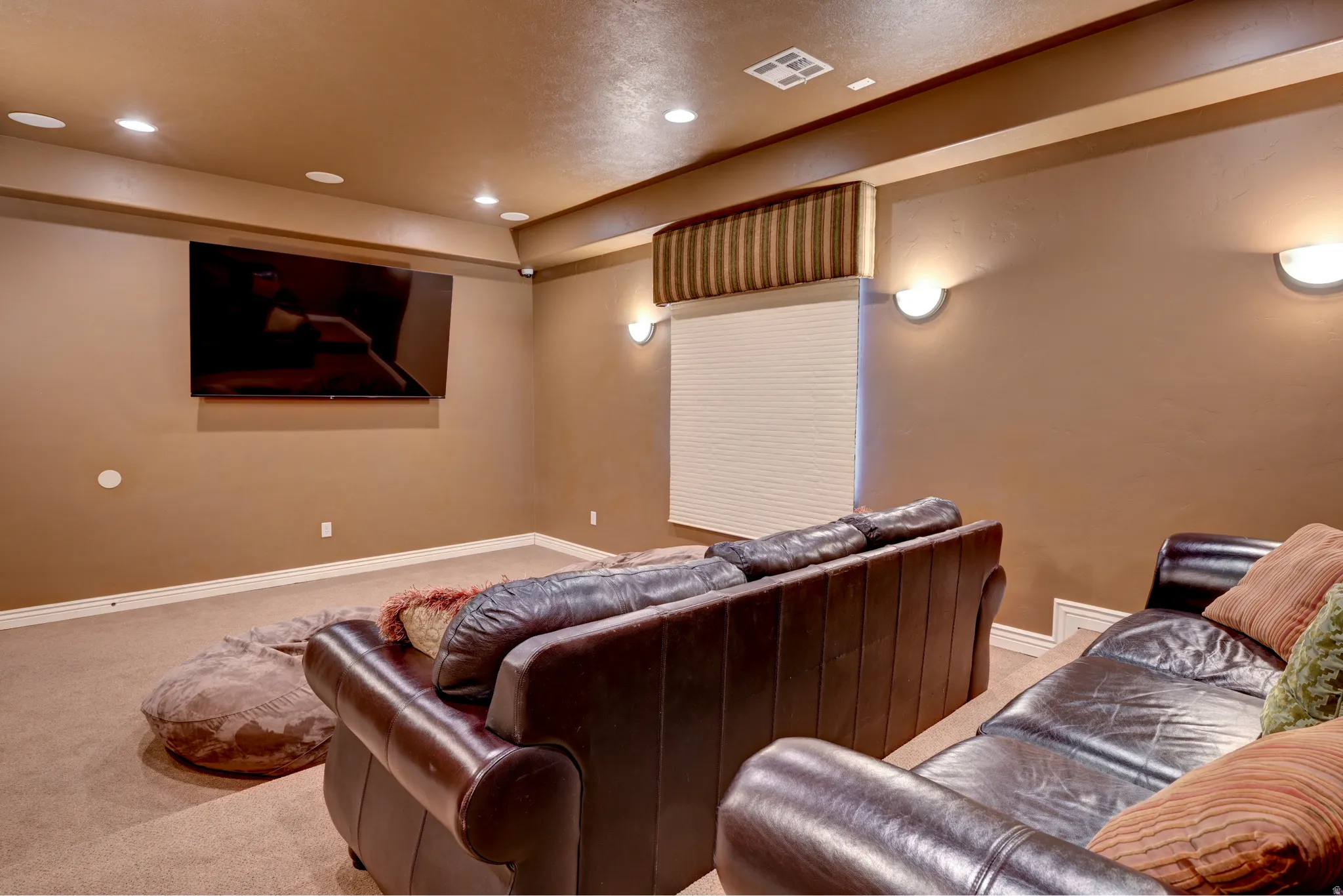 Spacious entertainment room featuring recessed ceiling lighting, wall-mounted sconces, a large wall-mounted screen, and neutral-toned carpeting