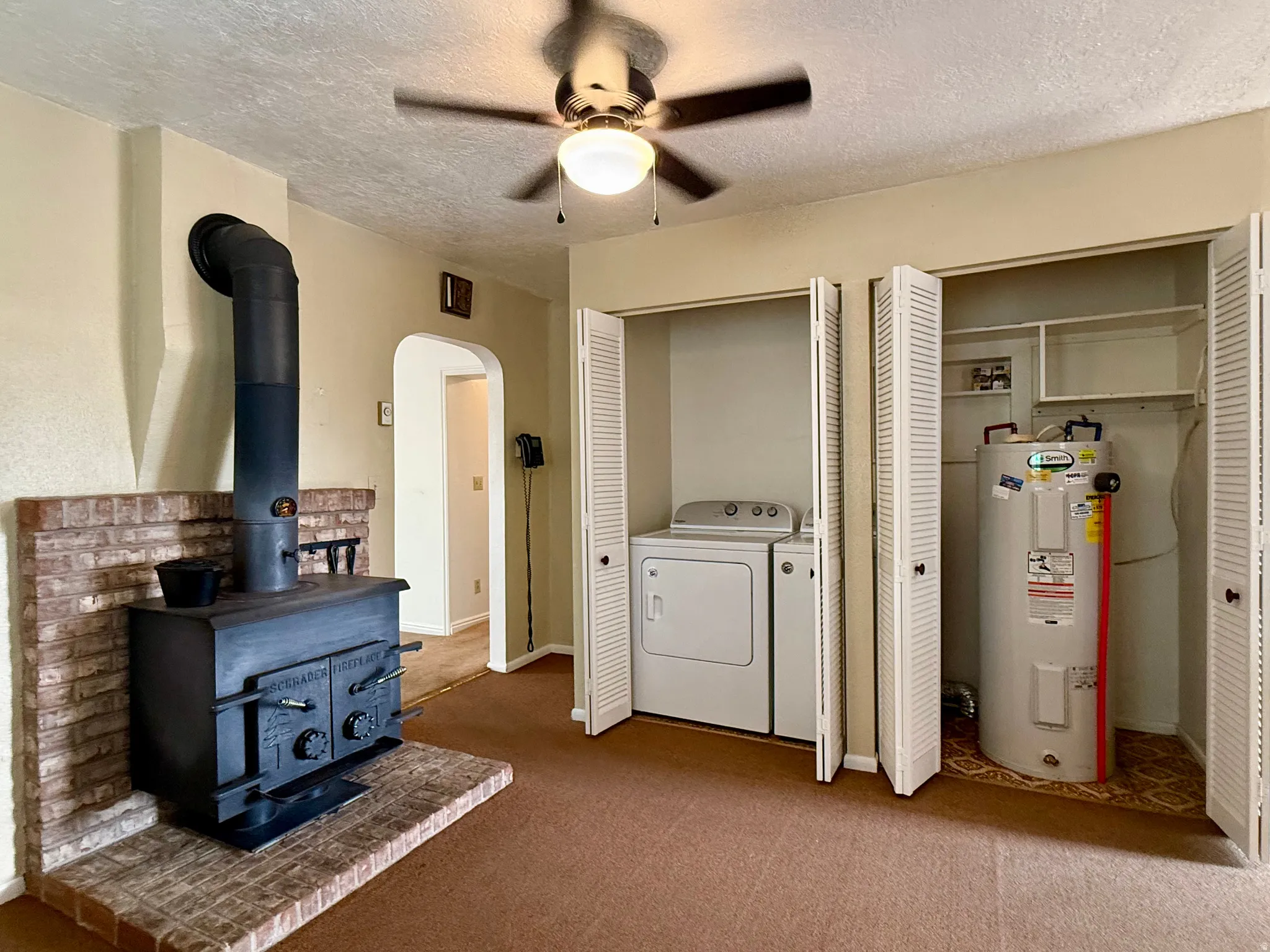 Brick hearth with a black wood-burning stove, brown carpet flooring, ceiling fan with integrated lighting, and bi-fold closet doors revealing a washer and dryer