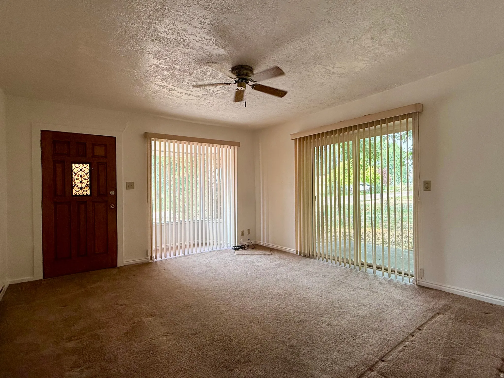 Expansive main living area featuring a warm brown carpet, a decorative wood entry door with an ornamental window insert, and vertical blinds on the window and sliding glass door