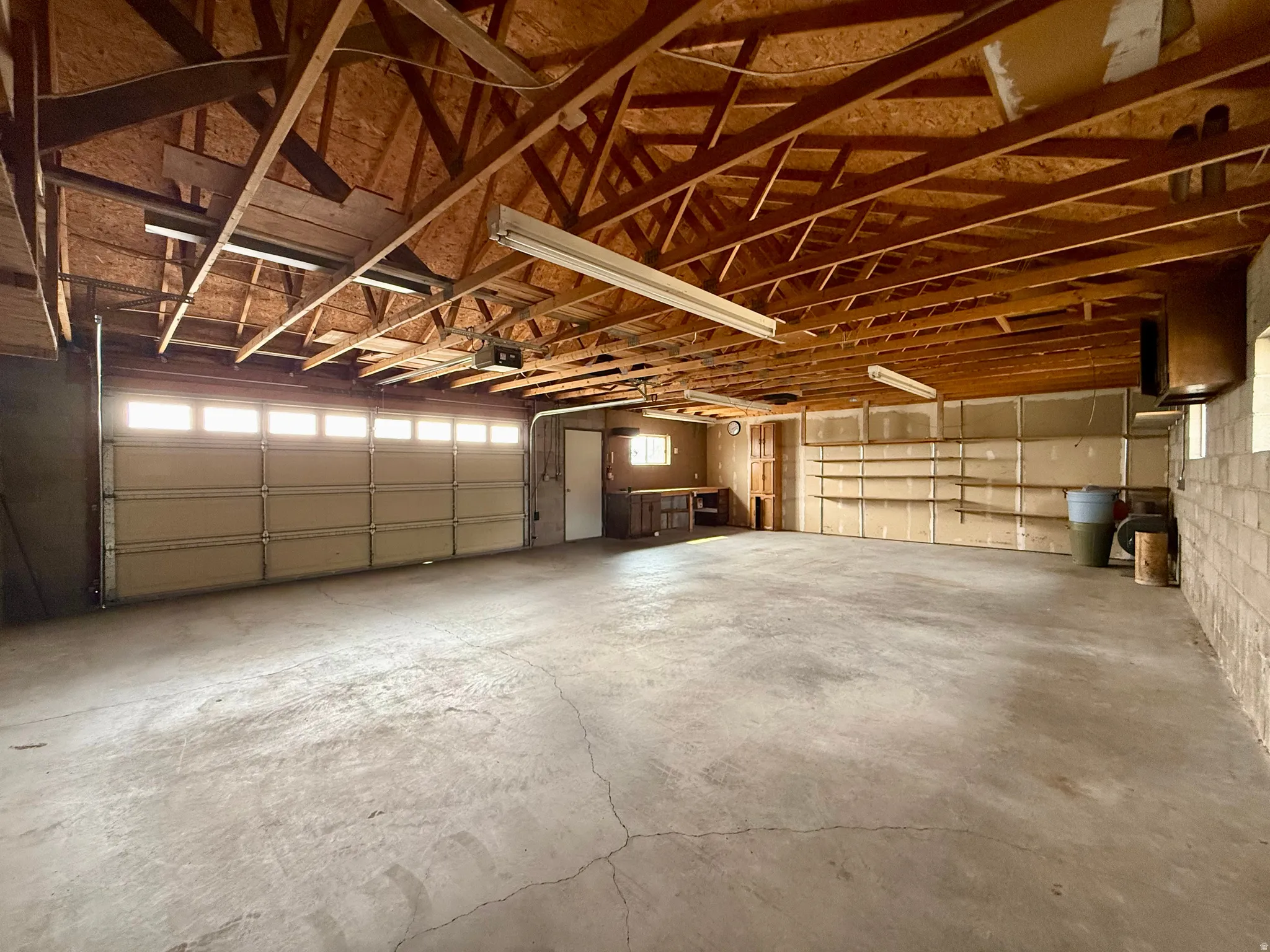Spacious garage featuring concrete flooring, exposed wood beam ceiling, two overhead garage doors with frosted window panels, built-in shelving, and a built-in workbench