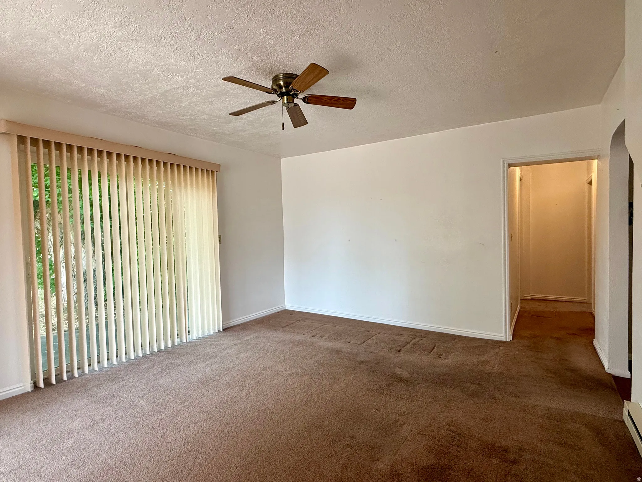 Living area featuring a sliding glass door with vertical blinds, brown carpeting, a ceiling fan with wood-finish blades, white painted walls, and a textured ceiling