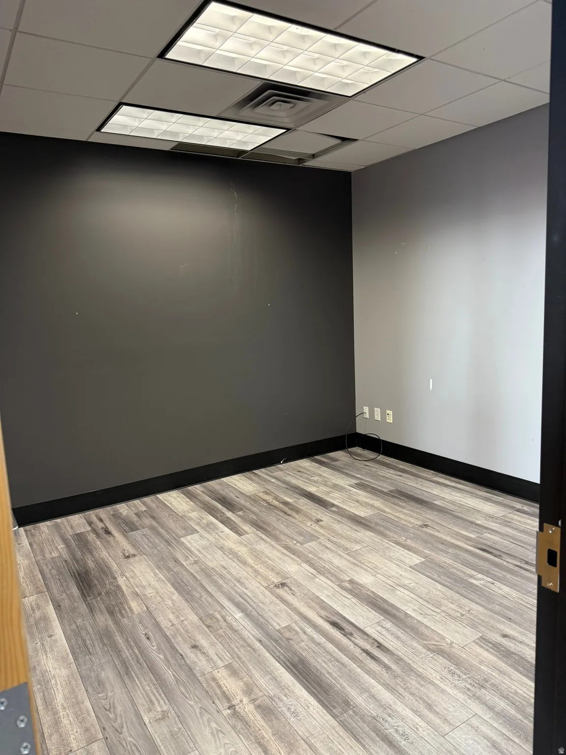 Room featuring wood-finish flooring, dark accent wall, suspended ceiling with recessed lighting, and black baseboards