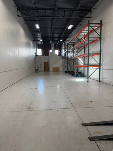 Spacious industrial interior featuring concrete flooring, high ceilings with exposed metal joists, multiple lighting fixtures, and integrated metal shelving units