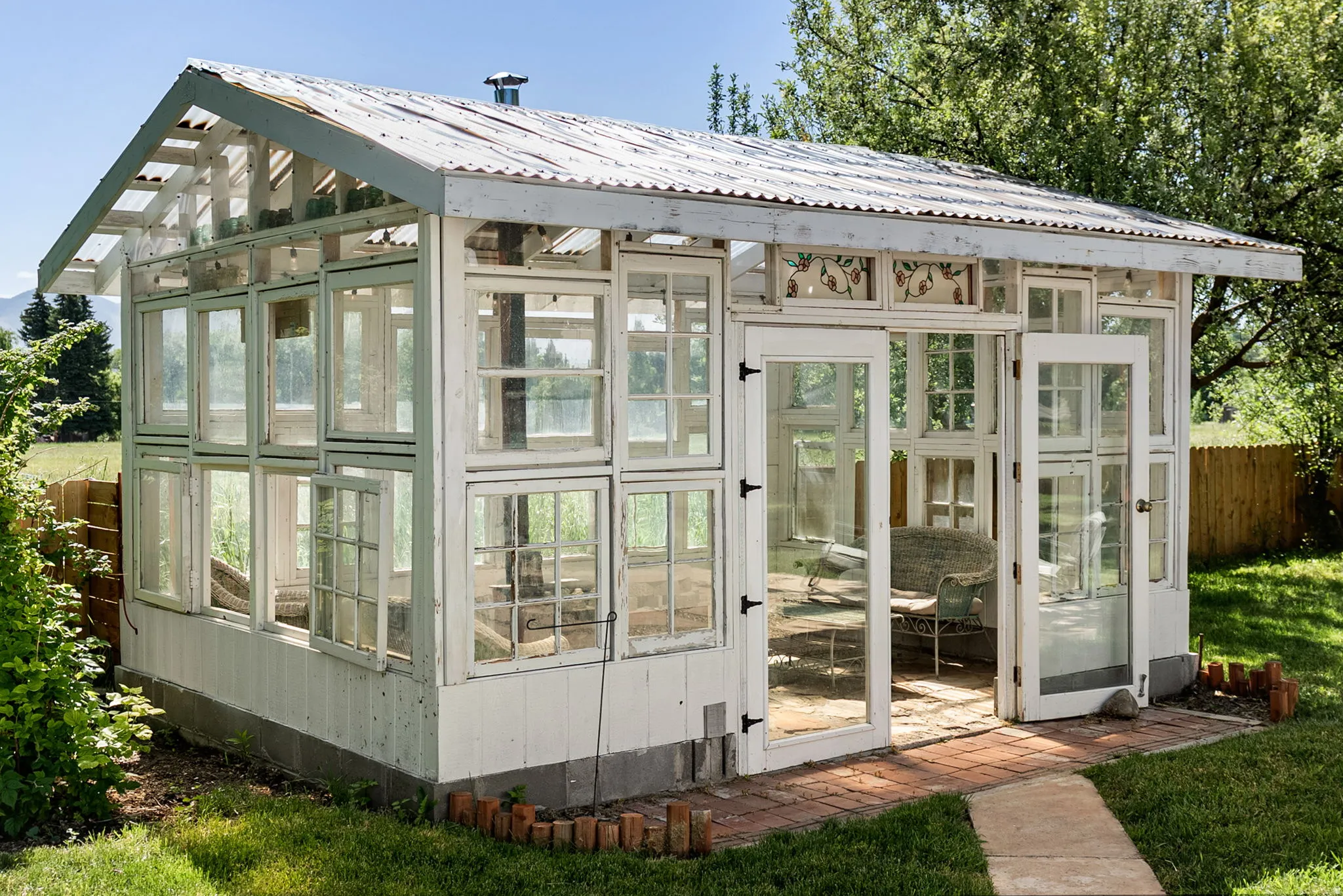 Detached greenhouse featuring a corrugated metal roof, multiple paneled windows, double French doors, and a brick paver entry