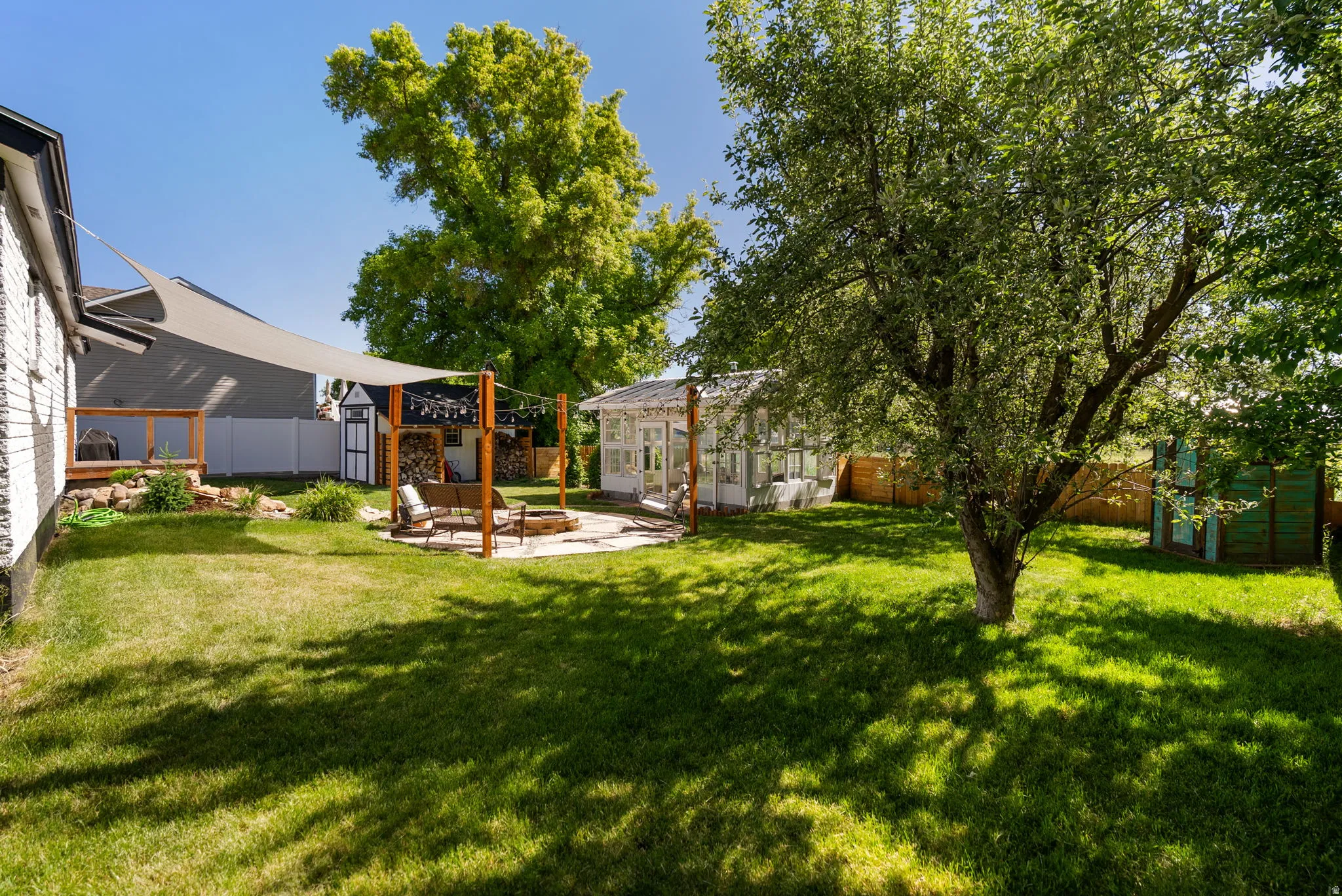 Expansive backyard featuring a manicured lawn, shade sail, paver patio with fire pit, and a greenhouse structure
