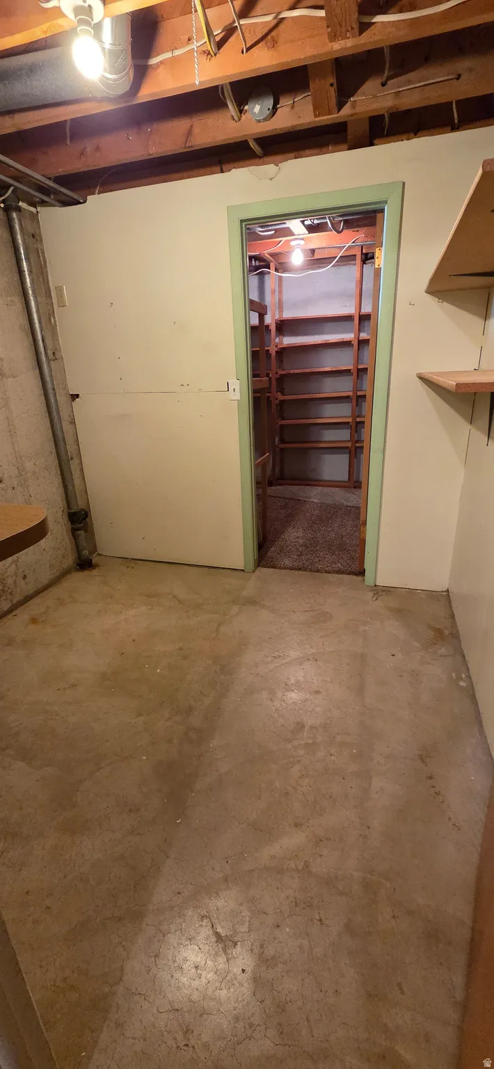 Utility room featuring exposed ceiling joists, concrete flooring, and a doorway to a storage area with built-in shelving