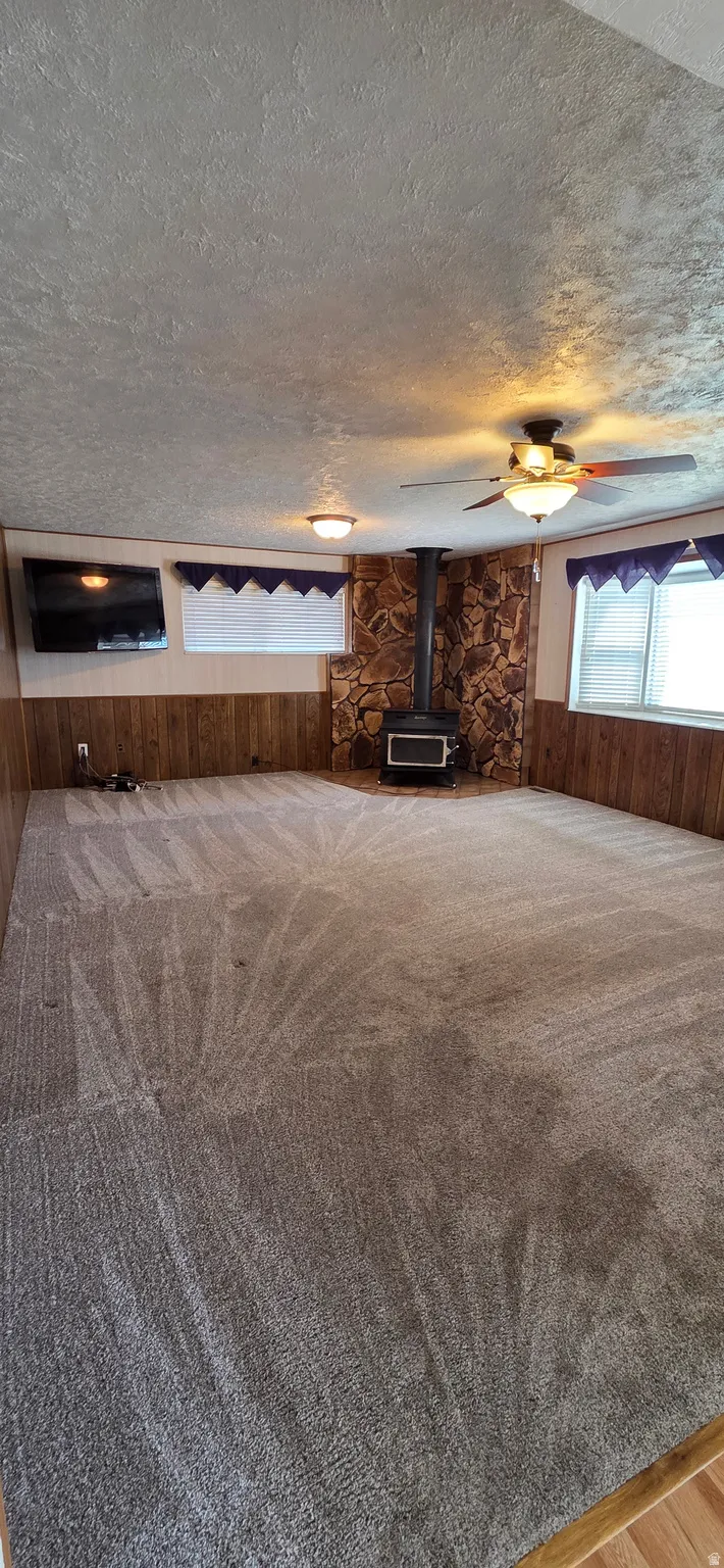 Carpeted room featuring a wood-burning stove with a stone surround, wood paneling, and a ceiling fan with integrated lighting