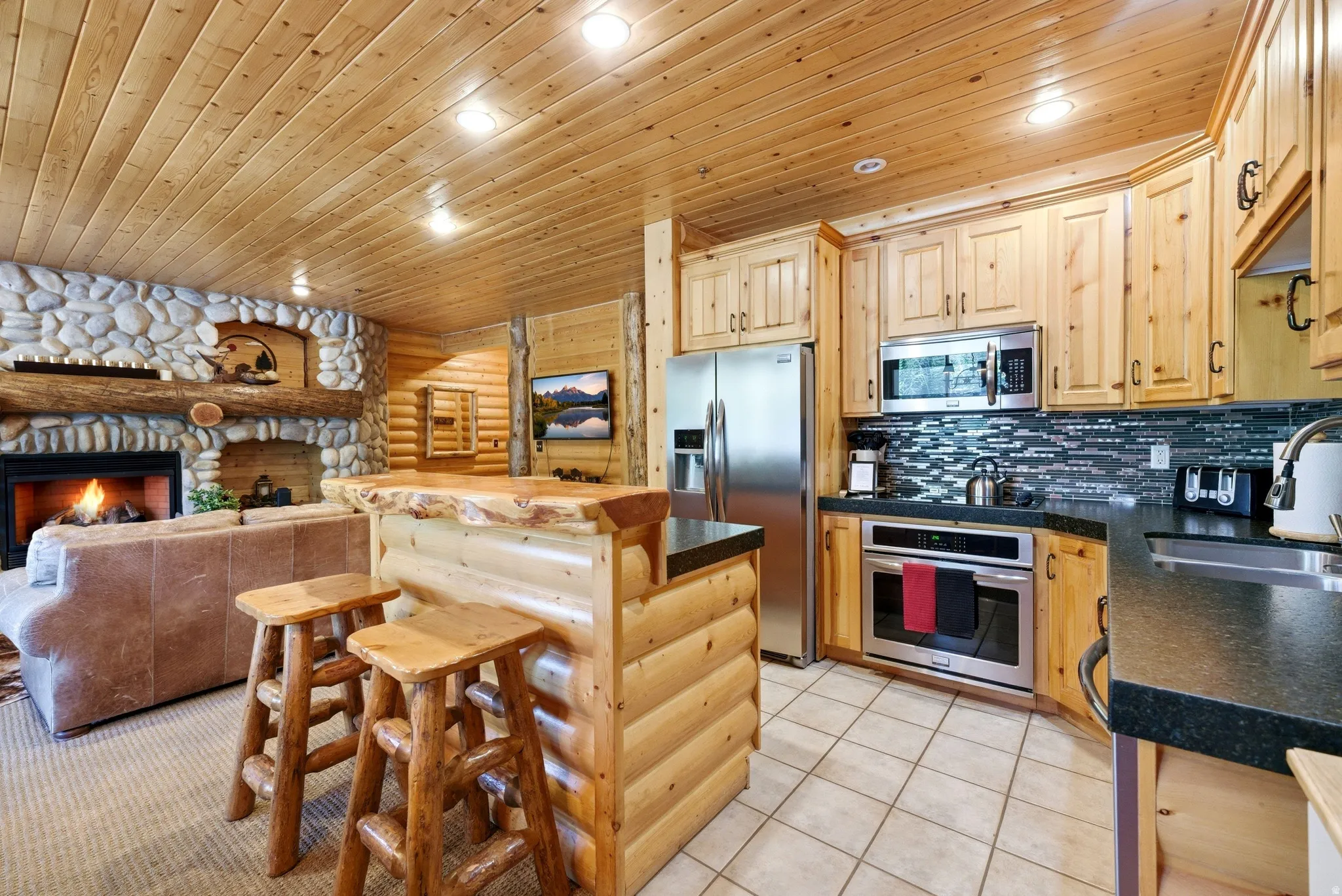 Kitchen and living area featuring knotty pine paneling, a stone fireplace, stainless steel appliances, and a wood-finish bar