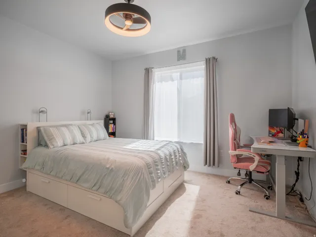 Bright bedroom featuring a large window with privacy sheers and drapery, neutral carpet flooring, a contemporary ceiling light fixture, and light gray wall tones