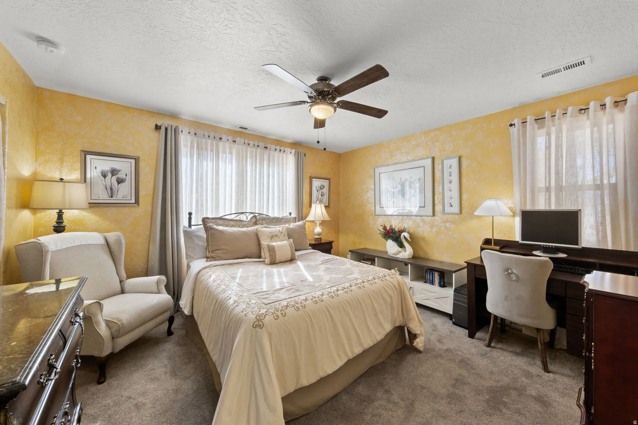 Carpeted room featuring textured walls, a ceiling fan with integrated lighting, and multiple windows with sheer and opaque drapery