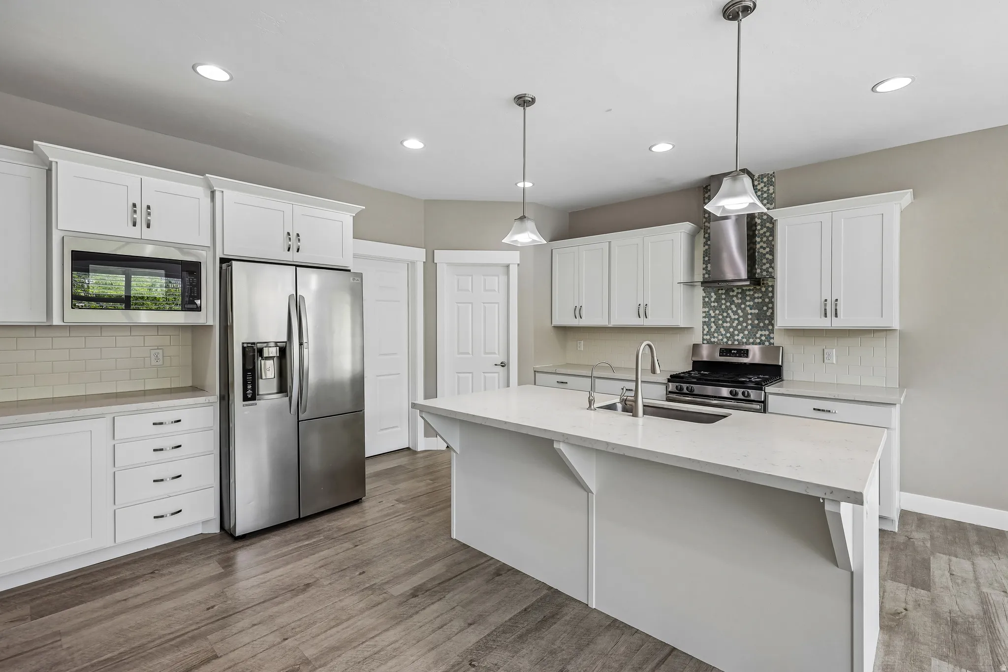 Modern kitchen featuring white shaker cabinetry, stainless steel appliances, a center island with sink and pendant lighting, wood-finish flooring, and a decorative tile backsplash