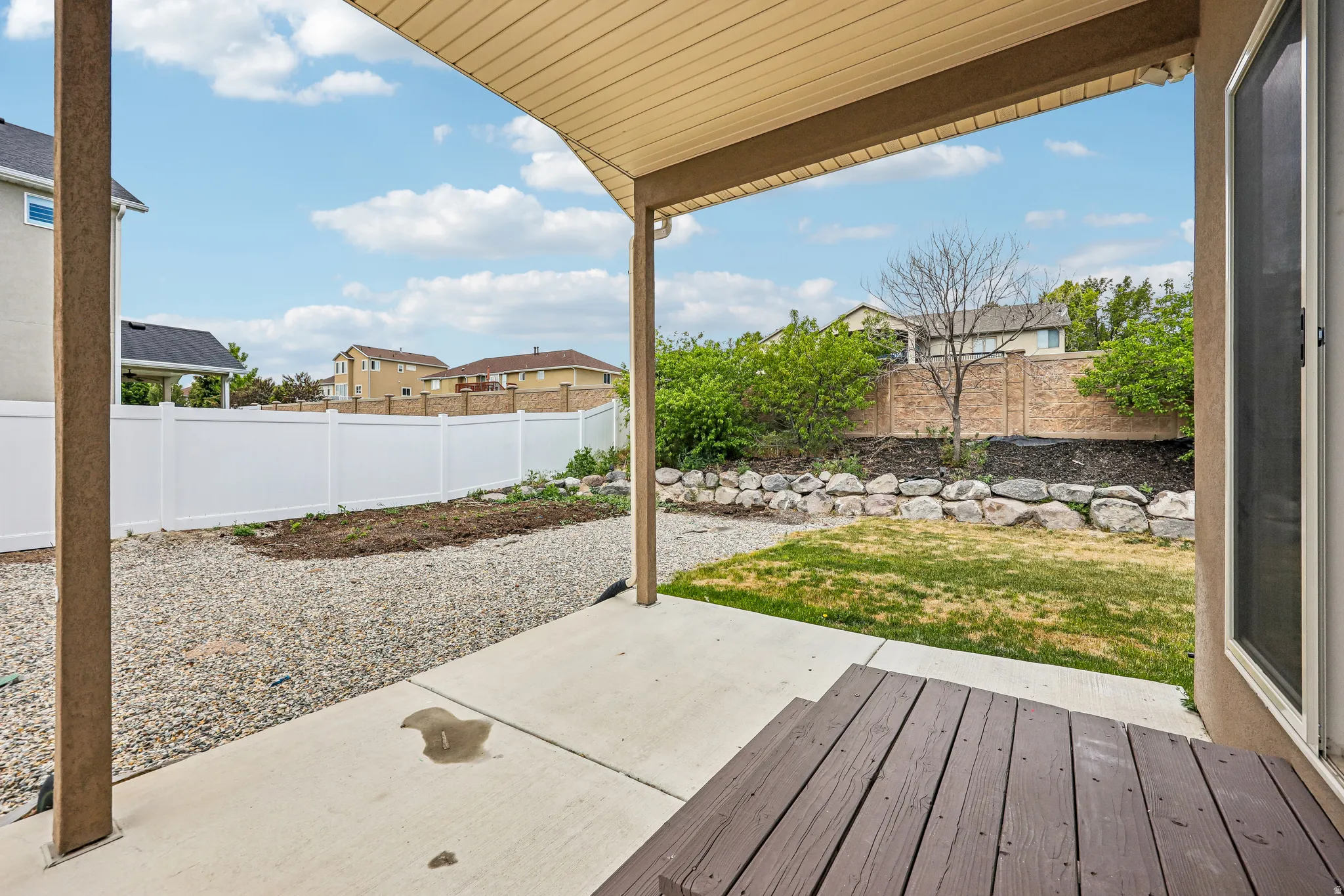 Covered patio with concrete flooring, leading to a gravel area, a retaining wall with rock features, and a grassy section