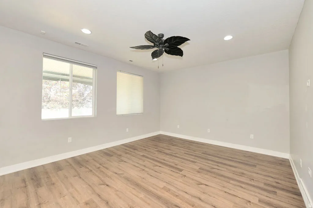 Spacious room featuring wood-finish flooring, light gray walls, two windows with blinds, a decorative ceiling fan, and recessed lighting