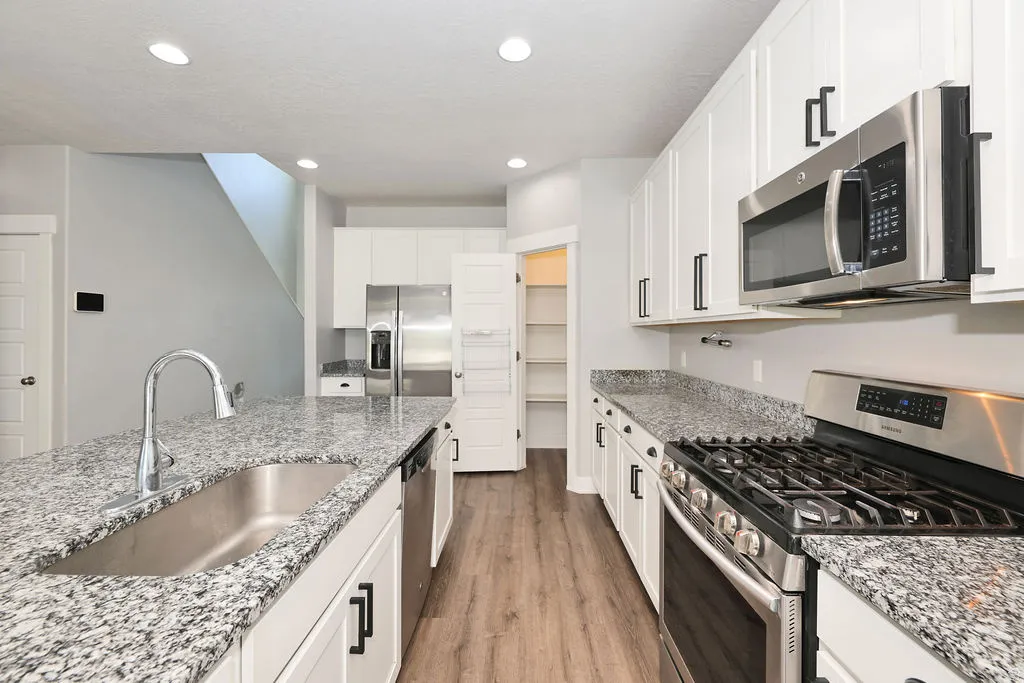 Contemporary kitchen featuring a spacious island with an undermount sink, granite-style countertops, stainless steel appliances, white shaker cabinetry with dark hardware, and wood-finish flooring