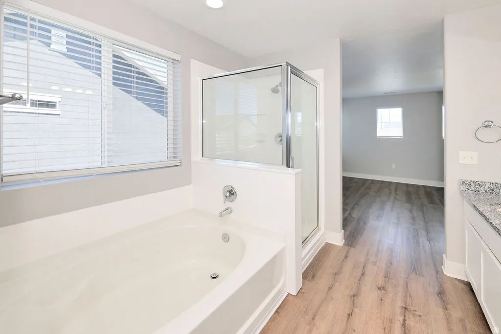 Bathroom featuring a built-in soaking tub, a glass-enclosed shower, wood-finish flooring, and a vanity with a speckled countertop