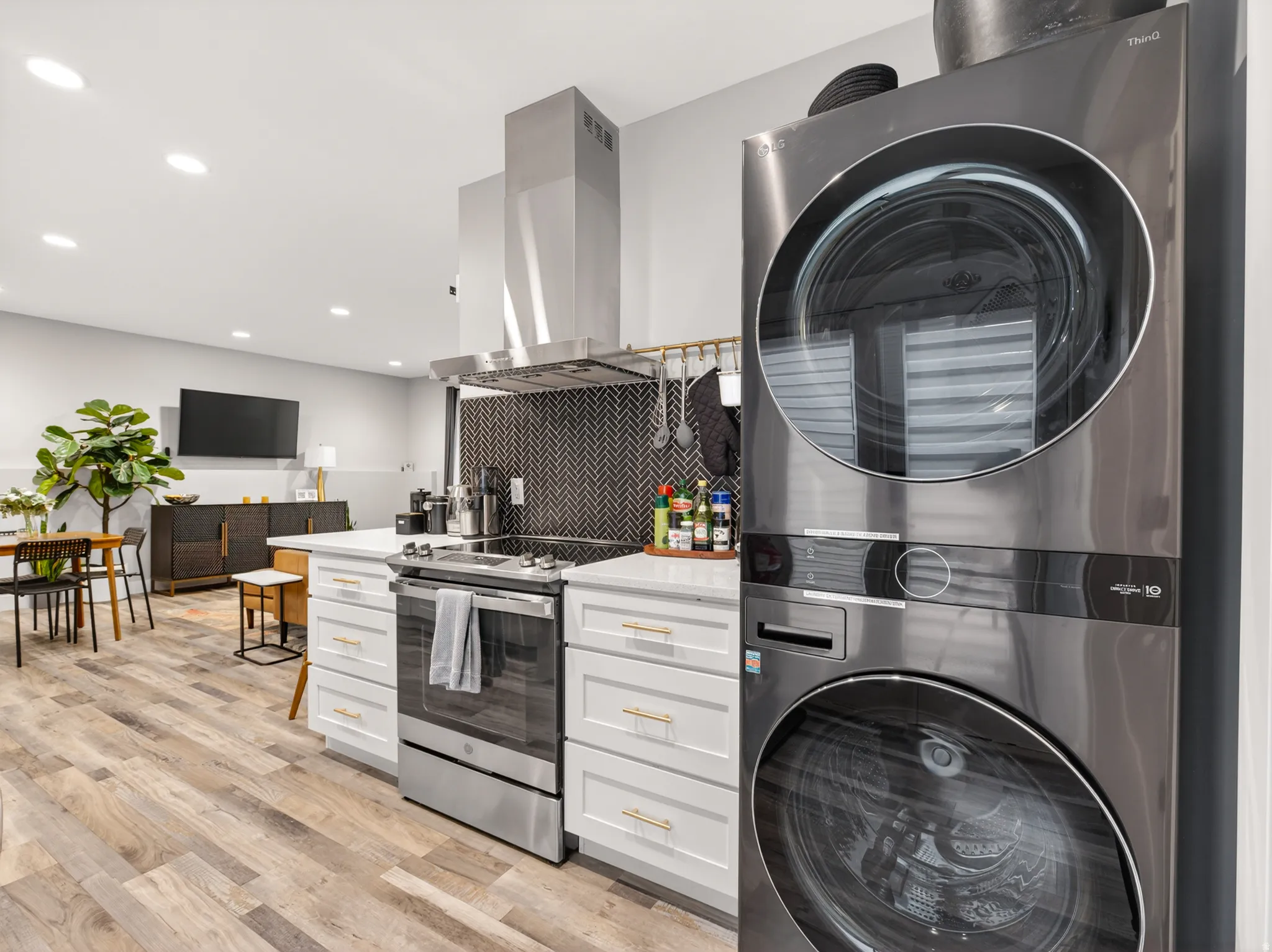 Integrated kitchen and laundry area featuring a stacked washer and dryer, stainless steel range with overhead vent, white cabinetry with gold-finish hardware, herringbone tile backsplash, and wood-finish flooring