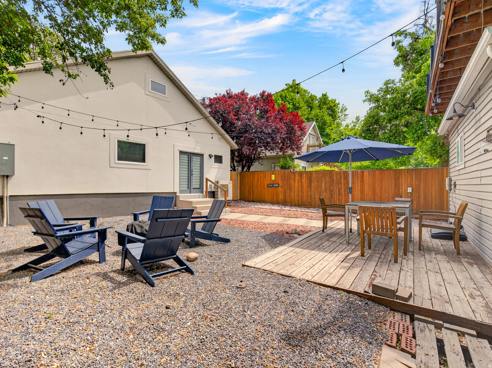 Spacious backyard featuring a gravel patio area with string lighting, a wooden privacy fence, a secondary wooden deck, a sliding glass door entry, and mature landscaping
