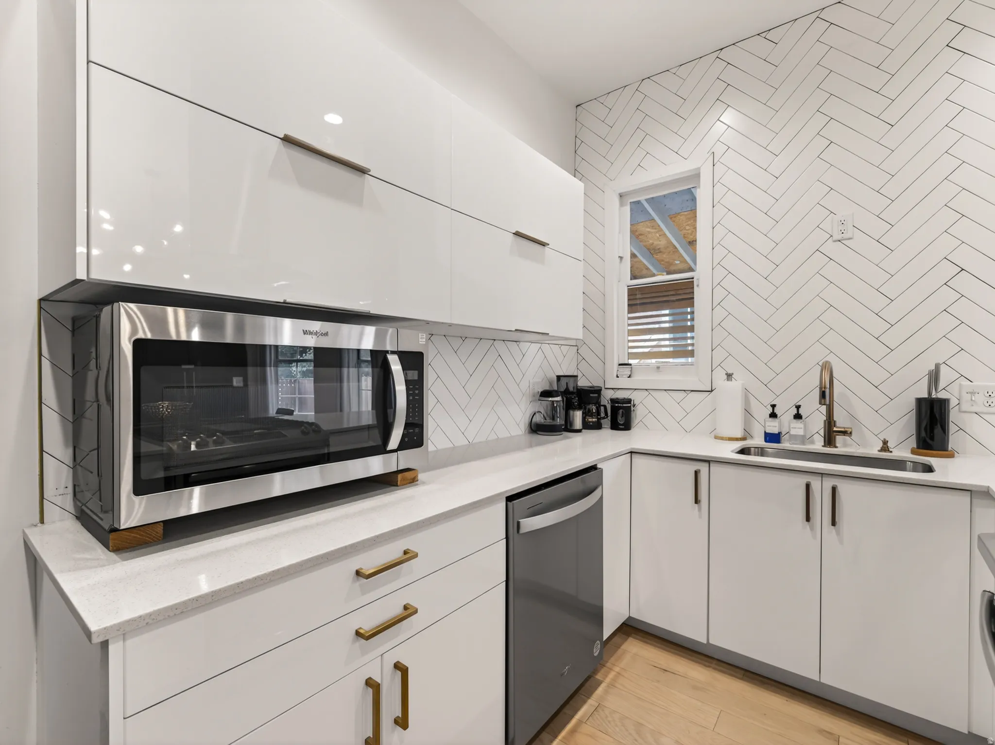 Modern kitchen featuring white high-gloss cabinetry with brass hardware, a herringbone tile backsplash, and light-toned wood-finish flooring