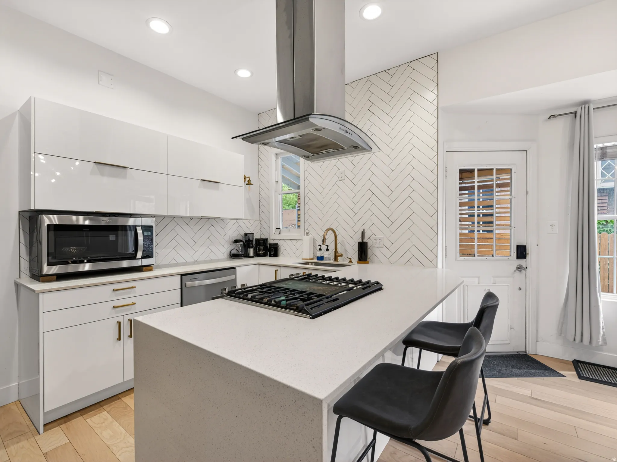 Modern kitchen featuring white high-gloss cabinetry, light wood-finish flooring, a white herringbone tile backsplash, and a large central island with a built-in gas cooktop
