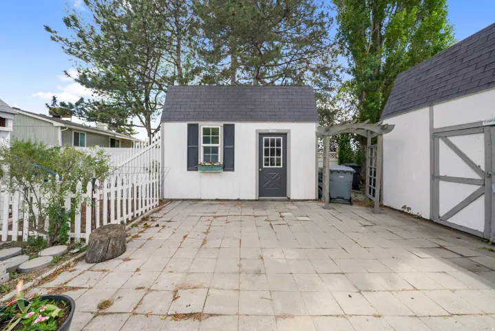 Paved patio area featuring two detached storage sheds, a white picket fence, and mature trees