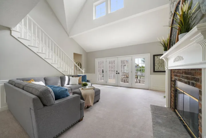 Spacious living area featuring a vaulted ceiling with upper windows, a carpeted floor, a traditional brick fireplace with a stone hearth and white mantel, and a staircase with white balusters