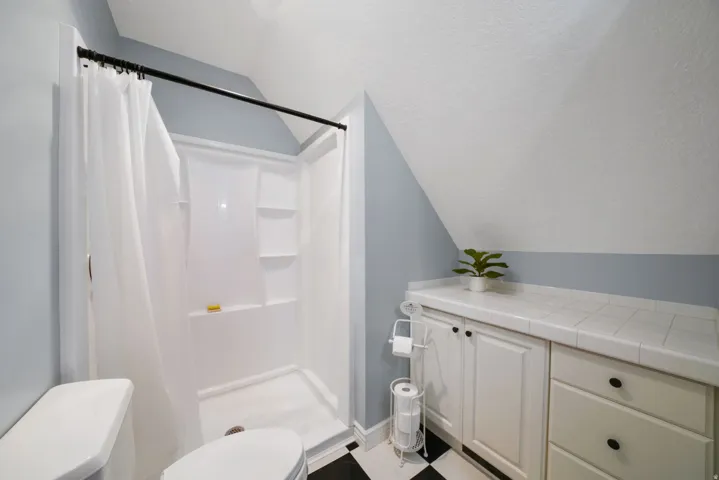 Bathroom featuring a white shower stall with built-in shelving, white tiled countertop, white cabinetry with black hardware, and black and white checkerboard flooring