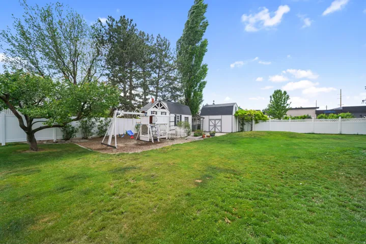 Expansive backyard featuring a lush lawn, white vinyl fencing, mature trees, and a functional storage shed