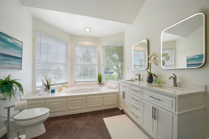 Spacious bathroom featuring a built-in soaking tub with surrounding bay windows, a dual vanity with white cabinetry and undermount sinks, square-framed mirrors, and dark tile flooring