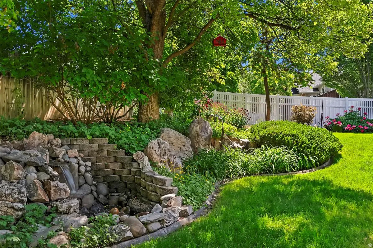 Landscaped backyard featuring a tiered stone waterfall, stone steps, mature trees, vibrant plantings, and a white picket fence