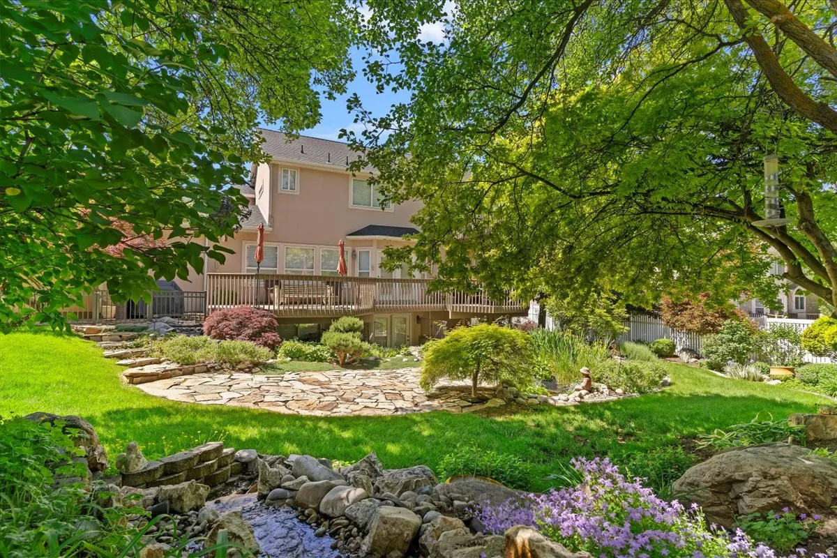 Lush green backyard featuring a multi-level wood deck, stone-paved patio, and a decorative rock-lined water feature