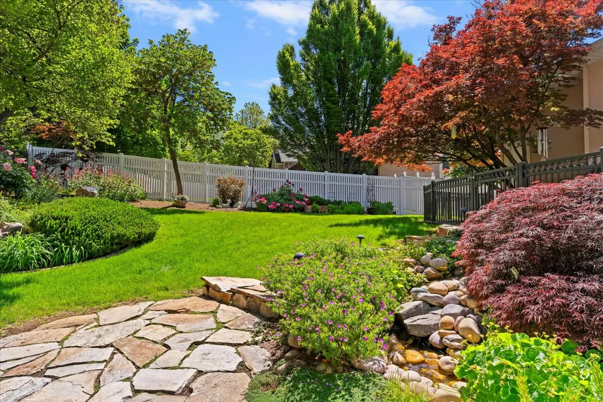 Landscaped backyard featuring a stone pathway, a manicured lawn, a white picket fence, a tiered rock garden with a water feature, and mature trees including a vibrant Japanese maple