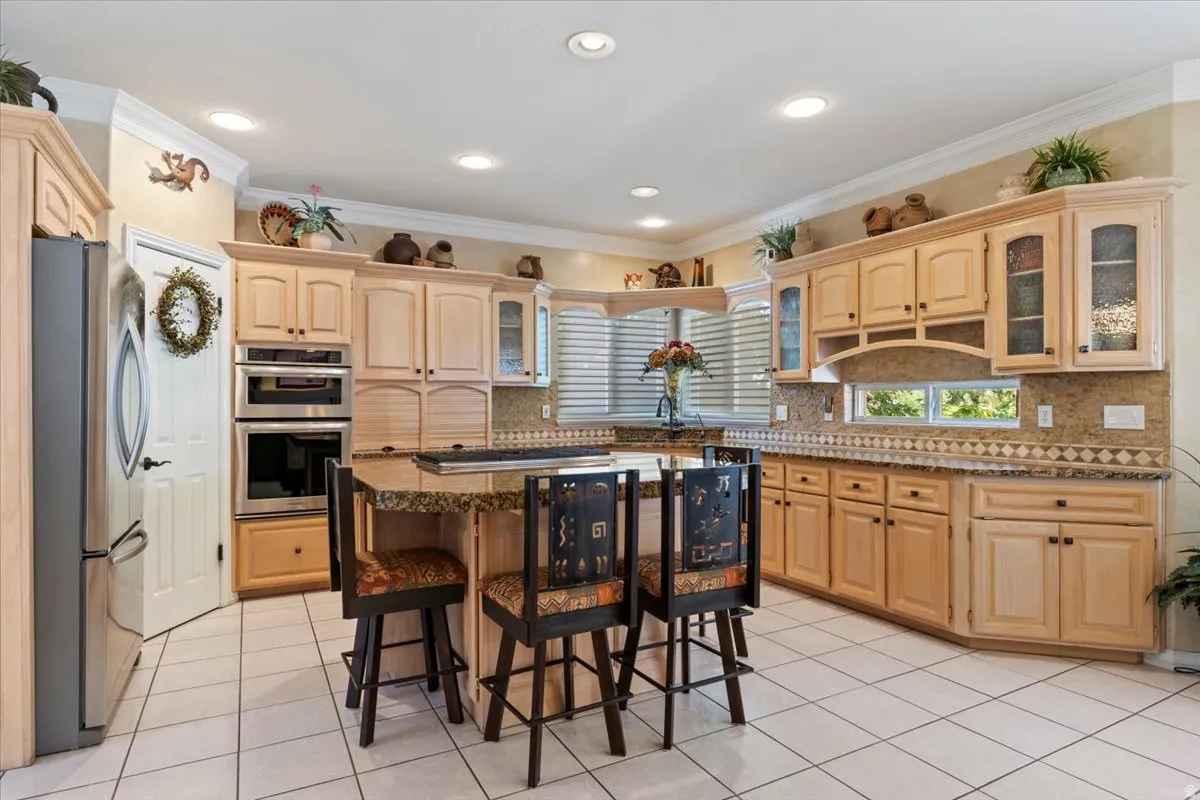 Kitchen featuring light wood cabinetry, granite countertops, tile backsplash, built-in double ovens, and a central island with bar seating
