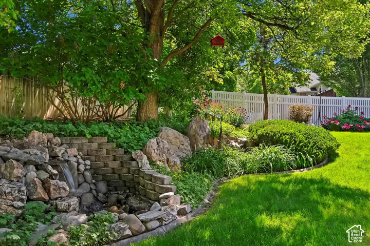 Landscaped backyard featuring a tiered stone waterfall, stone steps, mature trees, vibrant plantings, and a white picket fence